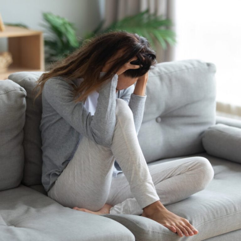 Stressful young mixed race woman sitting alone on sofa, holding head in hands, feeling desperate, suffering from relations break up or domestic violence. Unhealthy lady having strong headache.